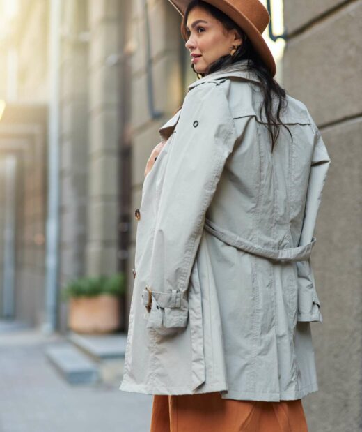 Rear view of a young attractive and stylish caucasian woman wearing grey coat and hat looking aside while standing on the city street, walking on warm autumn day. Fashion and beautiful people concept